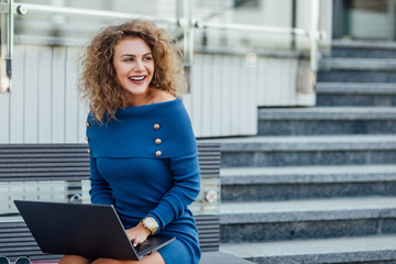 Young beautiful female with a laptop sits on a bench in the business part of the city. Young beautiful woman, freelancer, works on the laptop in summer wear blue dress. Lifestyle concept.