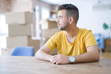 Young man sitting on the table with cardboard boxes behind him moving to new home looking to side, relax profile pose with natural face with confident smile.