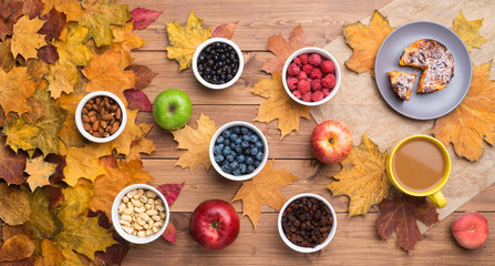 Seasonal autumn background. Frame of maple leaves and a cake, berries, raisins, apples, fruits, coffee and nuts over wooden background.