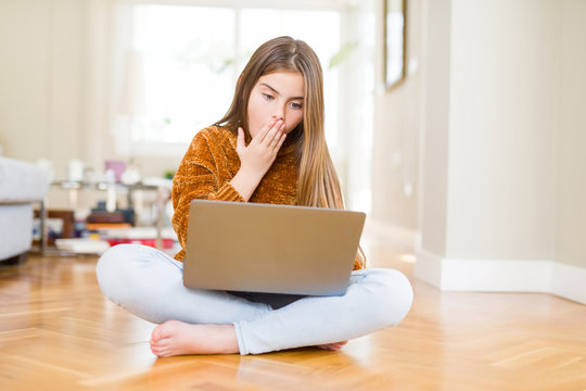 Beautiful young girl studying using laptop sitting on the floor at home cover mouth with hand shocked with shame for mistake, expression of fear, scared in silence, secret concept