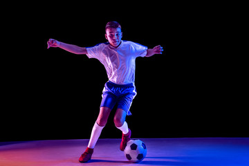 Fototapeta na wymiar Young boy as a soccer or football player in sportwear making a feint or a kick with the ball for a goal on dark studio background. Fit playing boy in action, movement, motion at game. Purple neon