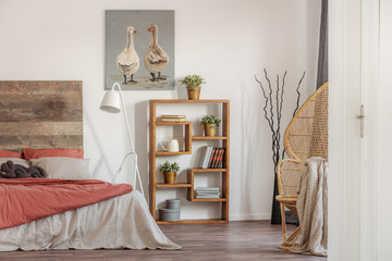 Real photo of a rustic bedroom interior with an oil painting, wooden bookcase and a bed with red and white sheets