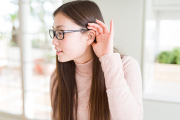 Beautiful Asian woman wearing glasses smiling with hand over ear listening an hearing to rumor or gossip. Deafness concept.