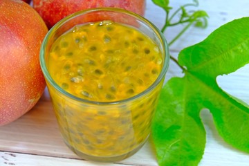 A glass of fresh passion fruits juice with green leaves and ripen passion fruits on white wooden table.
