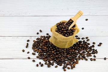 Top view of organic coffee beans in sack with a wooden spoon surrounded by roasted coffee beans on white wooden table. 