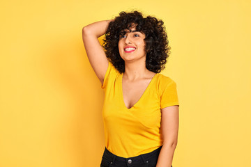 Young arab woman with curly hair wearing t-shirt standing over isolated yellow background smiling confident touching hair with hand up gesture, posing attractive and fashionable