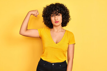 Young arab woman with curly hair wearing t-shirt standing over isolated yellow background Strong person showing arm muscle, confident and proud of power