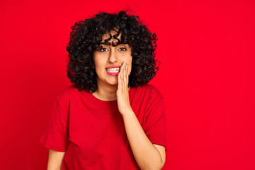 Young arab woman with curly hair wearing casual t-shirt over isolated red background touching mouth with hand with painful expression because of toothache or dental illness on teeth. Dentist concept.