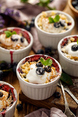 Blueberry crumble, stewed bilberries topped with crumble of flour, butter and sugar  with addition natural yogurt in a baking dishes on a wooden table, close-up. Homemade fruit dessert