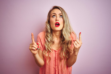 Young beautiful woman wearing t-shirt standing over pink isolated background amazed and surprised looking up and pointing with fingers and raised arms.
