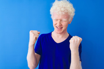 Young albino blond man wearing casual t-shirt standing over isolated blue background very happy and excited doing winner gesture with arms raised, smiling and screaming for success. 