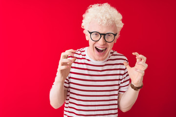 Young albino blond man wearing striped t-shirt and glasses over isolated red background celebrating crazy and amazed for success with arms raised and open eyes screaming excited. Winner concept