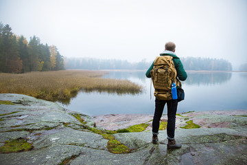 Mature man exploring Finland in the fall, looking into fog. Hiker with big backpack standing on mossy rock. Scandinavian landscape with misty sea and autumn forest. Back view