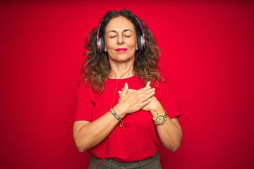 Middle age senior woman wearing headphones listening to music over red isolated background smiling with hands on chest with closed eyes and grateful gesture on face. Health concept.