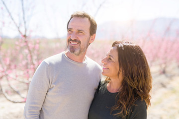 Fototapeta premium Beautiful middle age senior couple smiling in love at romantic garden of peach trees with pink petals on a sunny day of spring