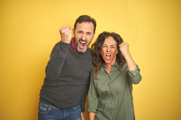 Beautiful middle age couple over isolated yellow background angry and mad raising fist frustrated and furious while shouting with anger. Rage and aggressive concept.