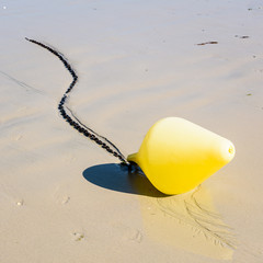 Close-up view of a large yellow buoy and its anchorage chain, used as a launching channel marker, lying on the wet sand on the beach in Penvenan, Brittany, France, under a bright sunshine.