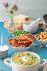Blue wooden table with bowls of thick green pea soup embellished with roasted rusks, green peas, slices of grilled bacon, crushed black pepper and cream.