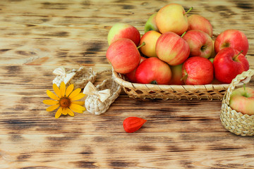 Red apples and autumn flowers lie in a wicker plate on a wooden table. Wicker bast shoes and apples...