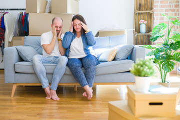 Young couple sitting on the sofa arround cardboard boxes moving to a new house with hand on head for pain in head because stress. Suffering migraine.