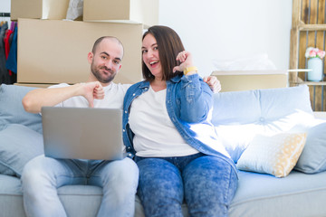 Young couple sitting on the sofa arround cardboard boxes moving to a new house using laptop with surprise face pointing finger to himself