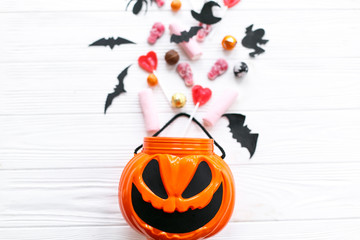 Halloween candy spilled from jack o lantern bucket with skulls, black bats, ghost, spider decorations on white wooden background, flat lay. Halloween sweets. Copy space. Trick or treat