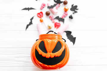 Halloween candy with skulls, black bats, ghost, spider decorations spilled from jack o lantern bucket on white wooden background, flat lay. Halloween sweets. Copy space. Trick or treat