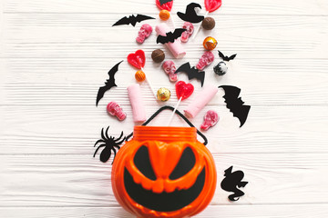 Happy Halloween. Halloween candy spilled from jack o lantern bucket with skulls, black bats, ghost, spider decorations on white wooden background, flat lay. Copy space. Trick or treat