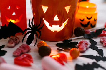 Halloween jack o lantern bucket, glowing candle, festive candy, skulls, black bats, ghost, spider decorations on white wooden background. Copy space. Trick or treat