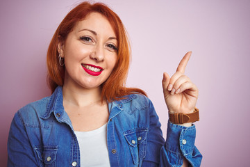 Youg beautiful redhead woman wearing denim shirt standing over isolated pink background with a big smile on face, pointing with hand and finger to the side looking at the camera.