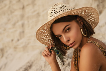 Outdoor portrait of young beautiful woman with luxury tanned skin wearing straw hat, snakeskin print swimwear, hoop earrings, posing in sand desert, at sunset. Copy, empty space for text