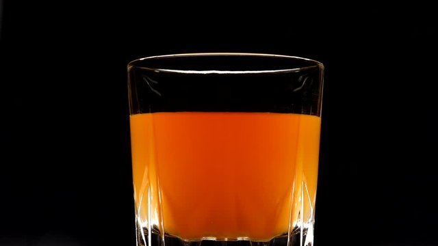 Rotating glass with yellow-orange sweet multifruit juice. Glass with juice on a dark background in backlight.