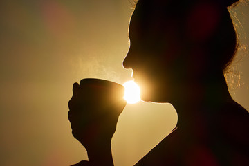 Young woman is enjoying cup of coffee outside with beautiful view of mountains and woods during a sunset/sunrise