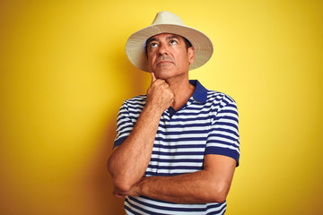 Handsome middle age man wearing striped polo and hat over isolated yellow background with hand on chin thinking about question, pensive expression. Smiling with thoughtful face. Doubt concept.