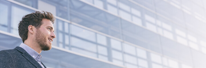Fototapeta premium Businessman looking away at city office building background panoramic banner. Young business man in his 30s happy thinking of his future at his job, career prospects.