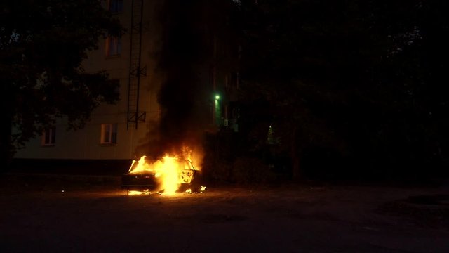 Auto on fire burning near a multi-storey building, slow motion. Fire alarm lights flashing on house. Theme of criminal revenge and set fire to someone else car. Automobile engulfed in flames of fire.