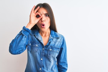 Young beautiful woman wearing casual denim shirt standing over isolated white background doing ok gesture shocked with surprised face, eye looking through fingers. Unbelieving expression.