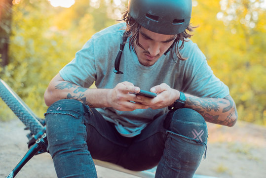 young cyclist guy , with tattoos, sitting on a bicycle in a protective helmet, uses the Internet in a smartphone, on a warm natural background. active lifestyle