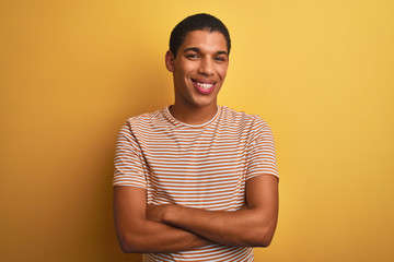 Young handsome arab man wearing striped t-shirt standing over isolated yellow background happy face smiling with crossed arms looking at the camera. Positive person.