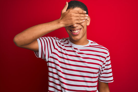 Young handsome arab man wearing striped t-shirt over isolated red background smiling and laughing with hand on face covering eyes for surprise. Blind concept.