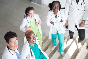 Doctors are sitting on the steps of the hospital. A group of young people of different gender, mixed race, in medical clothes, with phonendoscopes. Use smartphone, study medical records, study x-rays