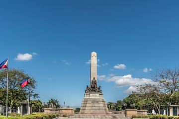 Manila, Philippines - March 5, 2019: Wide landscape shot of Obelisk with bronze statues of Jose Rizal stands on brown stone pedestal under blue sky in Rizal Park. Flags and green foliage.
