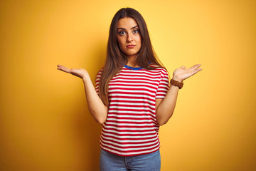 Young beautiful woman wearing striped t-shirt standing over isolated yellow background clueless and confused expression with arms and hands raised. Doubt concept.