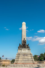 Manila, Philippines - March 5, 2019: Obelisk with bronze statues of Jose Rizal stands on brown stone pedestal under blue sky in Rizal Park. Flags and green foliage in back.
