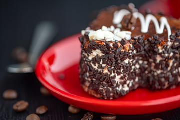 A delicious heart-shaped cake sprinkled with pieces of chocolate lies on a red plate. Photographed close-up on a wooden background.