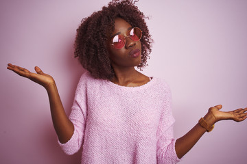Young african afro woman wearing sweater and sunglasses over isolated pink background clueless and...