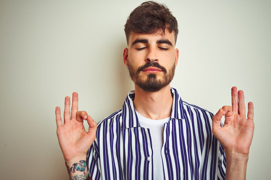 Young man with tattoo wearing striped shirt standing over isolated white background relax and smiling with eyes closed doing meditation gesture with fingers. Yoga concept.