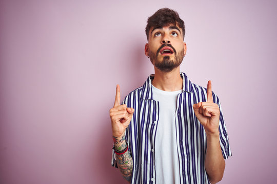 Young man with tattoo wearing striped shirt standing over isolated pink background amazed and surprised looking up and pointing with fingers and raised arms.