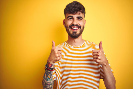 Young man with tattoo wearing striped t-shirt standing over isolated yellow background success sign doing positive gesture with hand, thumbs up smiling and happy. Cheerful expression 