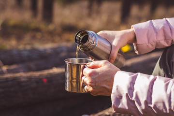 Woman pouring hot drink out of thermos into metal cup in autumn forest